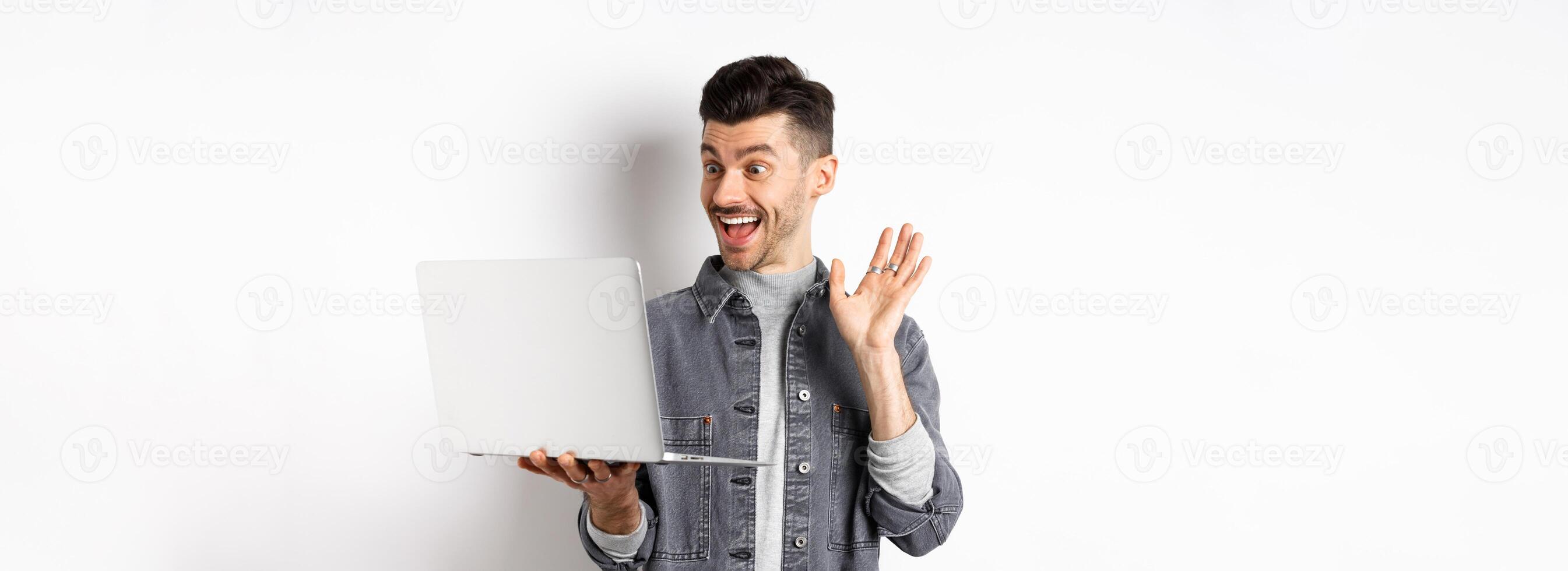Excited man waiving hand at laptop, chatting on computer and smiling friendly, standing on white background photo