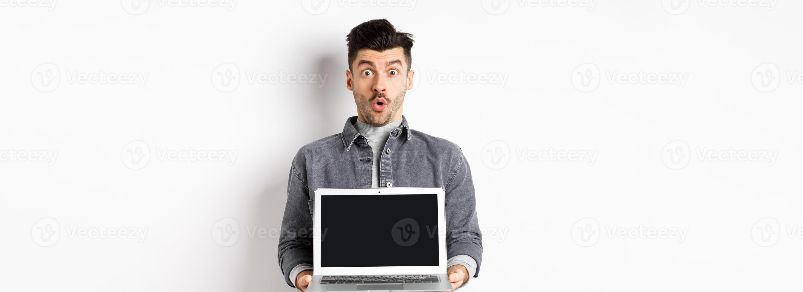 Excited man demostrated empty laptop screen, say wow and look at camera amazed, checking out online offer, showing webpage, standing on white background photo