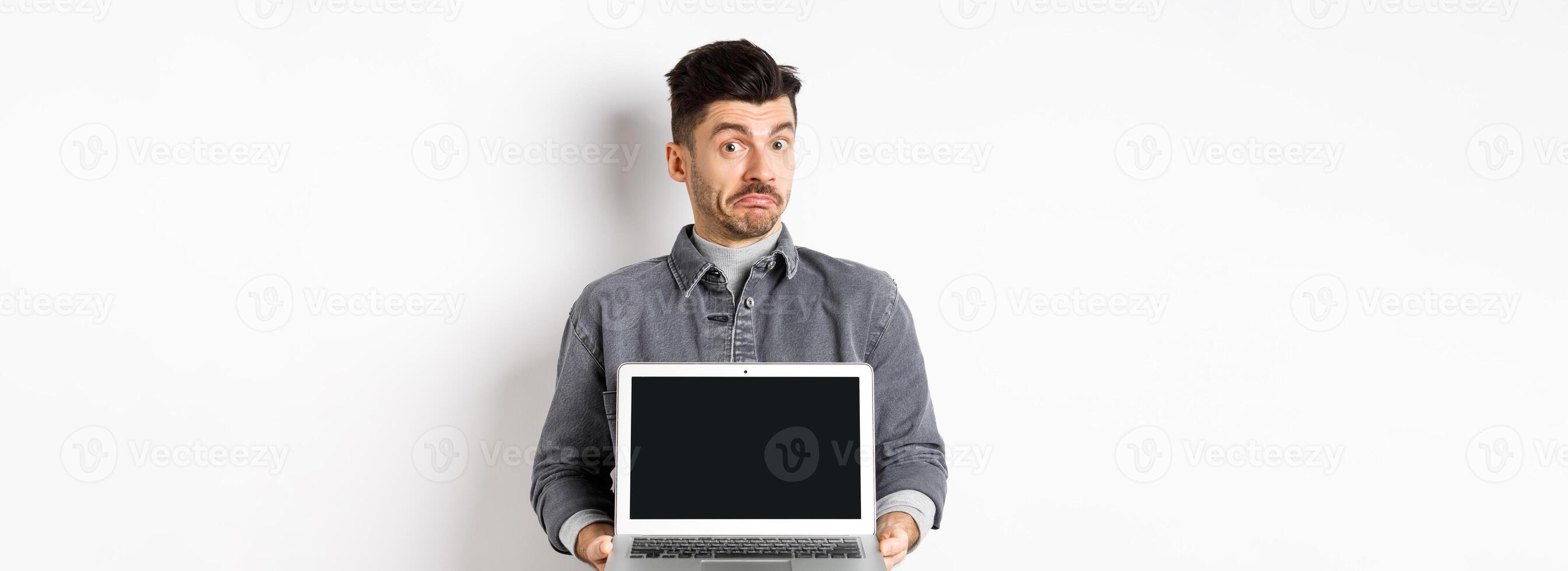 Indecisive young man pouting and shrugging while showing empty laptop screen, standing clueless on white background photo