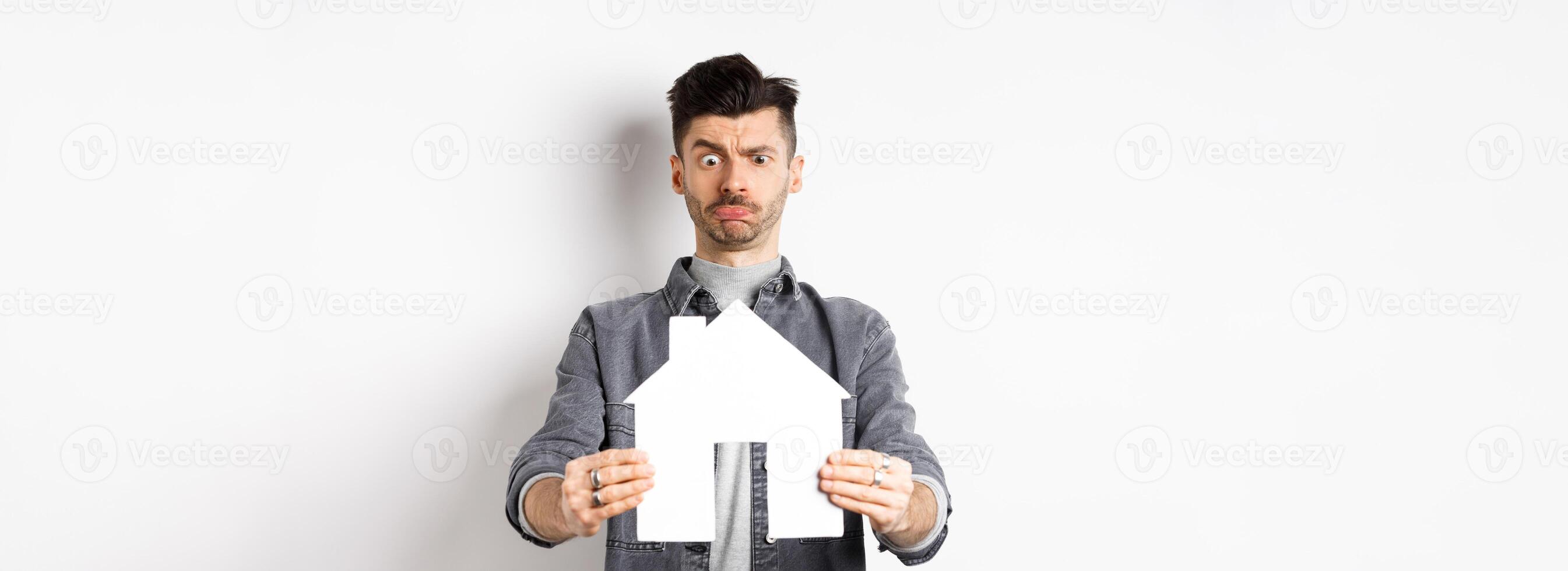 Real estate and insurance concept. Confused guy look at paper house cutout unsure, frowning perplexed, standing on white background photo