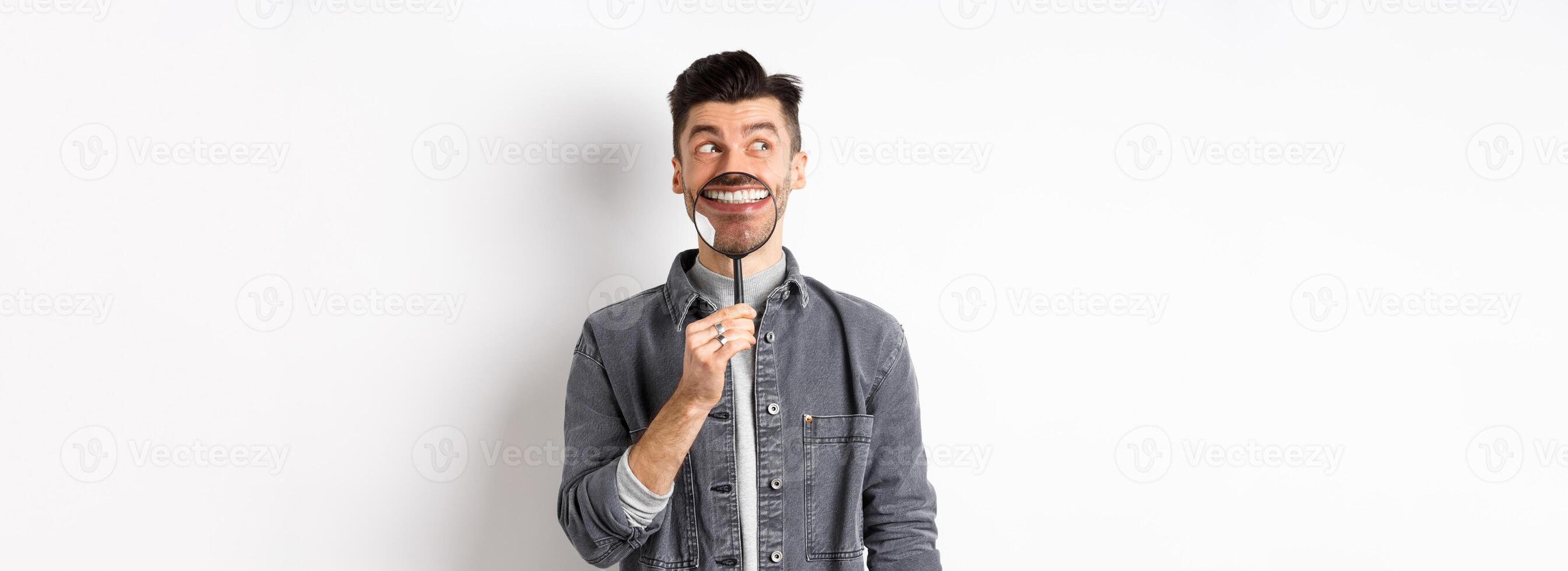 Handsome positive guy showing white perfect smile with magnifying glass, looking left at logo, standing against white background photo