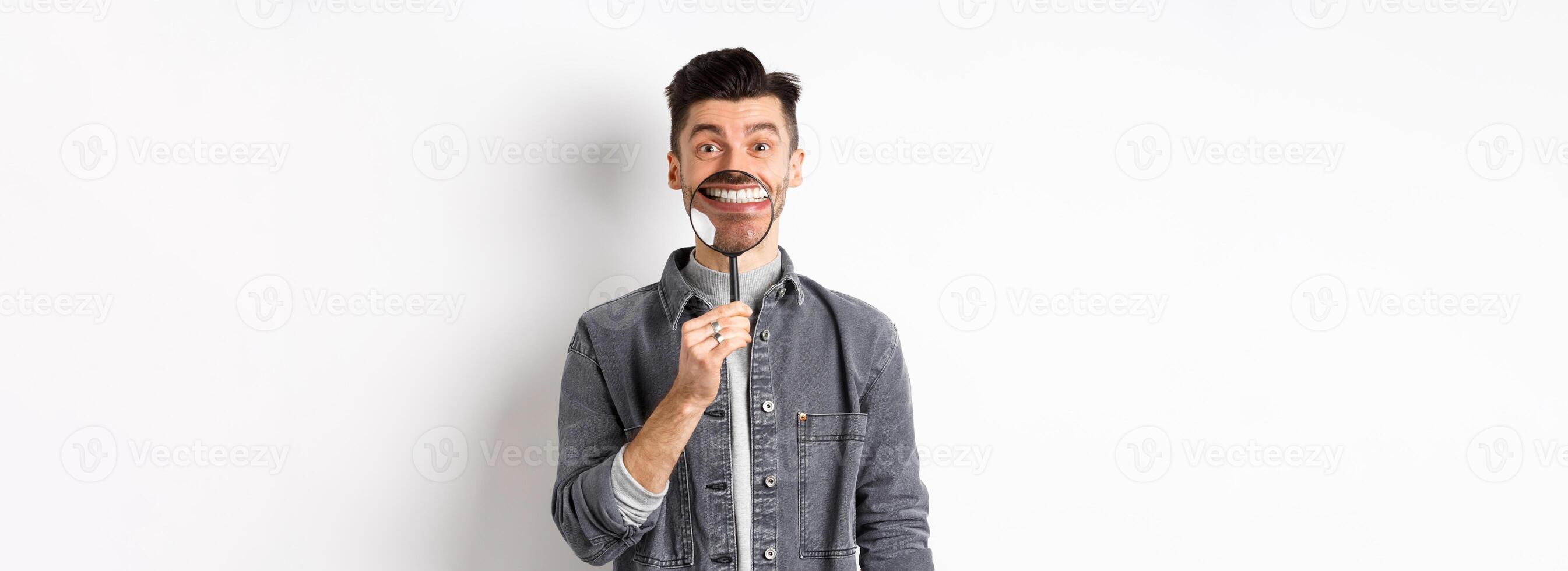 Handsome positive guy showing white perfect smile with magnifying glass, standing against white background photo