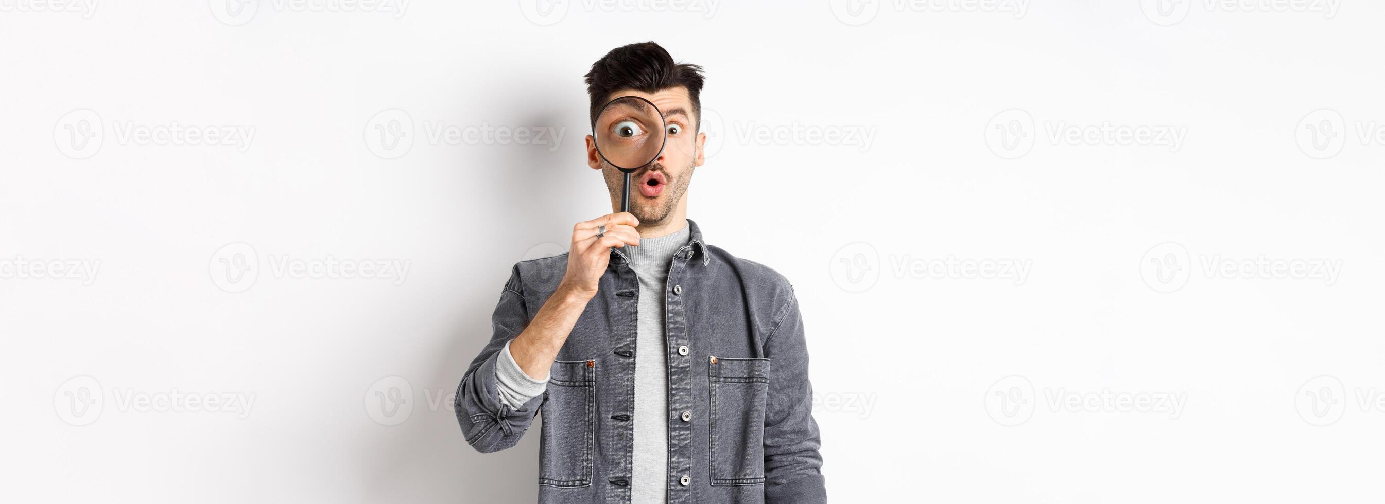 Image of excited guy look through magnifying glass, gasping amazed, big eye staring at interesting thing, found cool promo, standing on white background photo