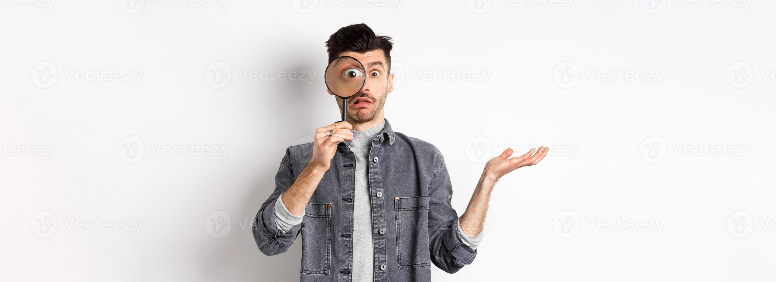 Confused man look through magnifying glass and shrugging clueless, standing on white background photo