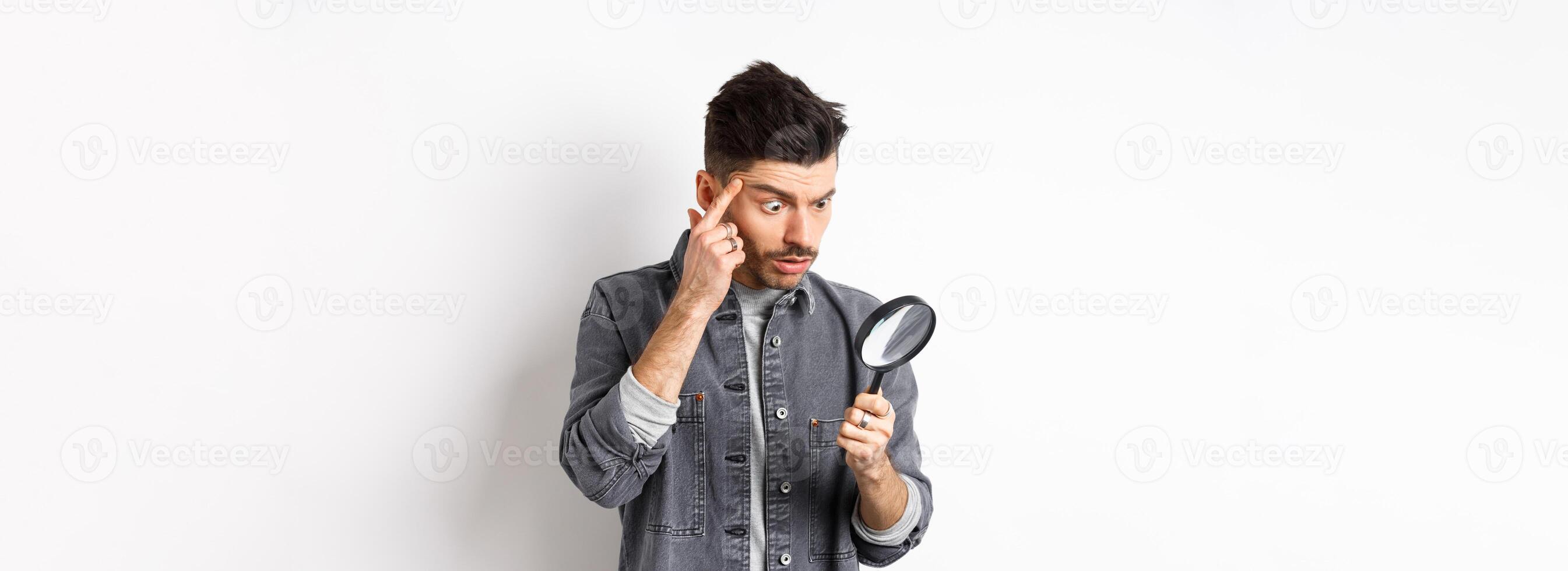 Man looking through magnifying glass and stretching eyelid to see clearly, standing on white background photo