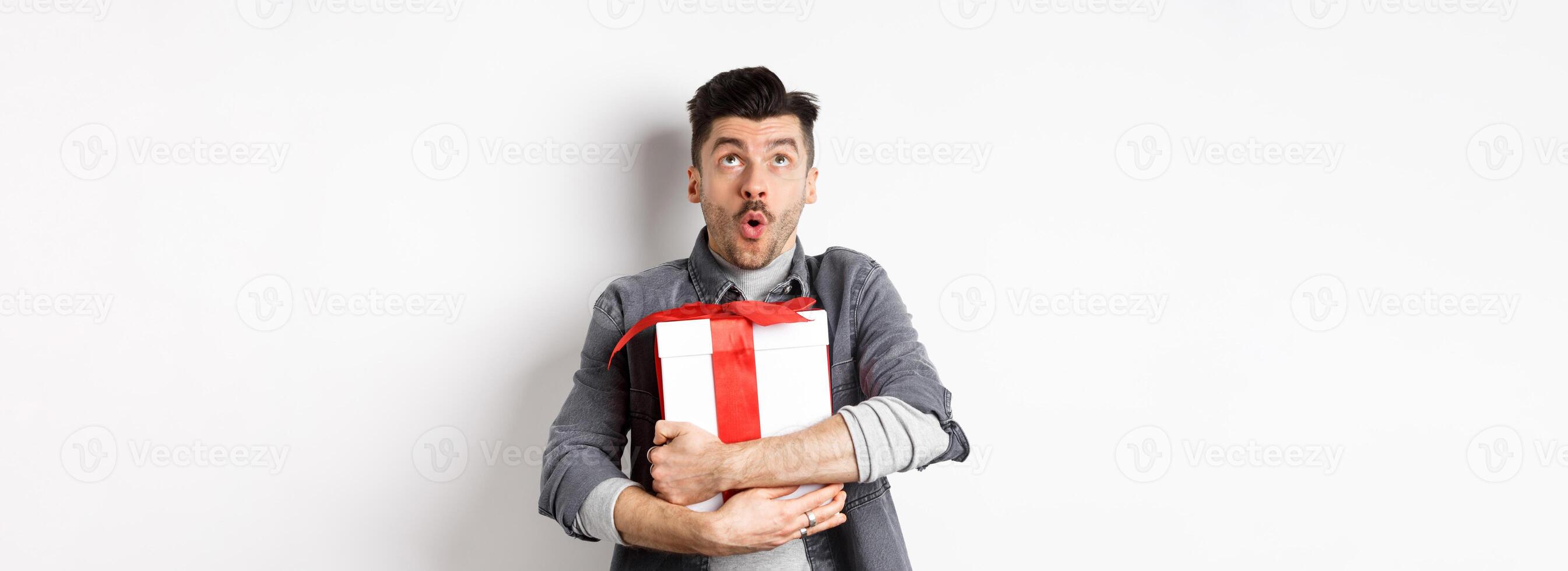 Surprised funny guy hugging big holiday gift and looking up, say wow amazed, checking out Valentines day special offer, standing on white background photo