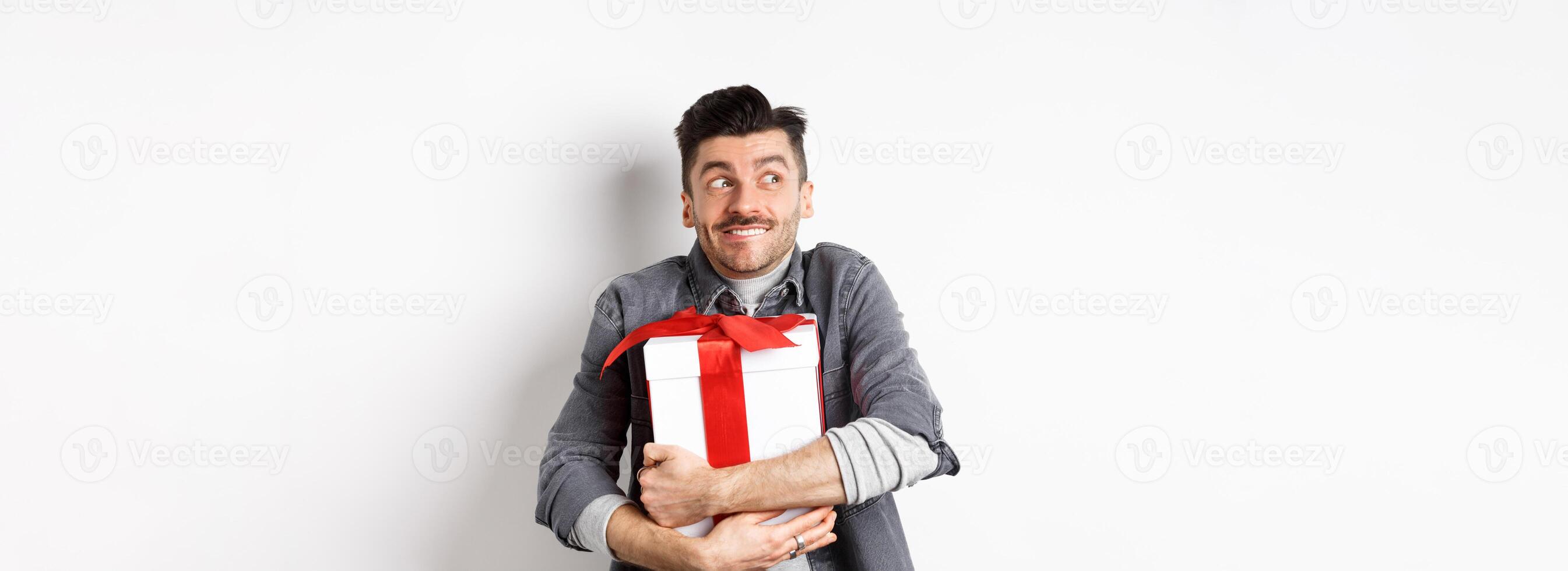 Happy boyfriend hugging gift from lover, looking aside at empty space and smiling excited, making romantic present on valentines day, standing on white background photo