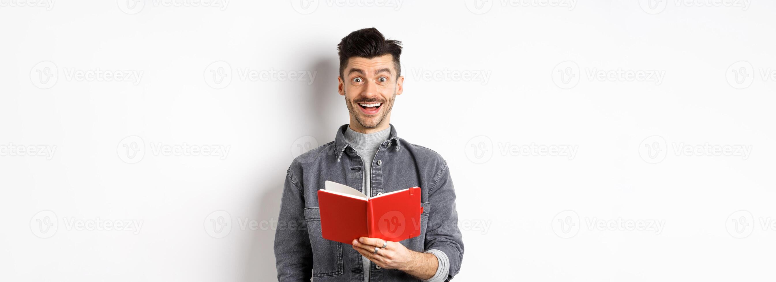Hobbies and leisure. Happy young man reading planner, holding diary or red journal and smiling, making notes, standing on white background photo