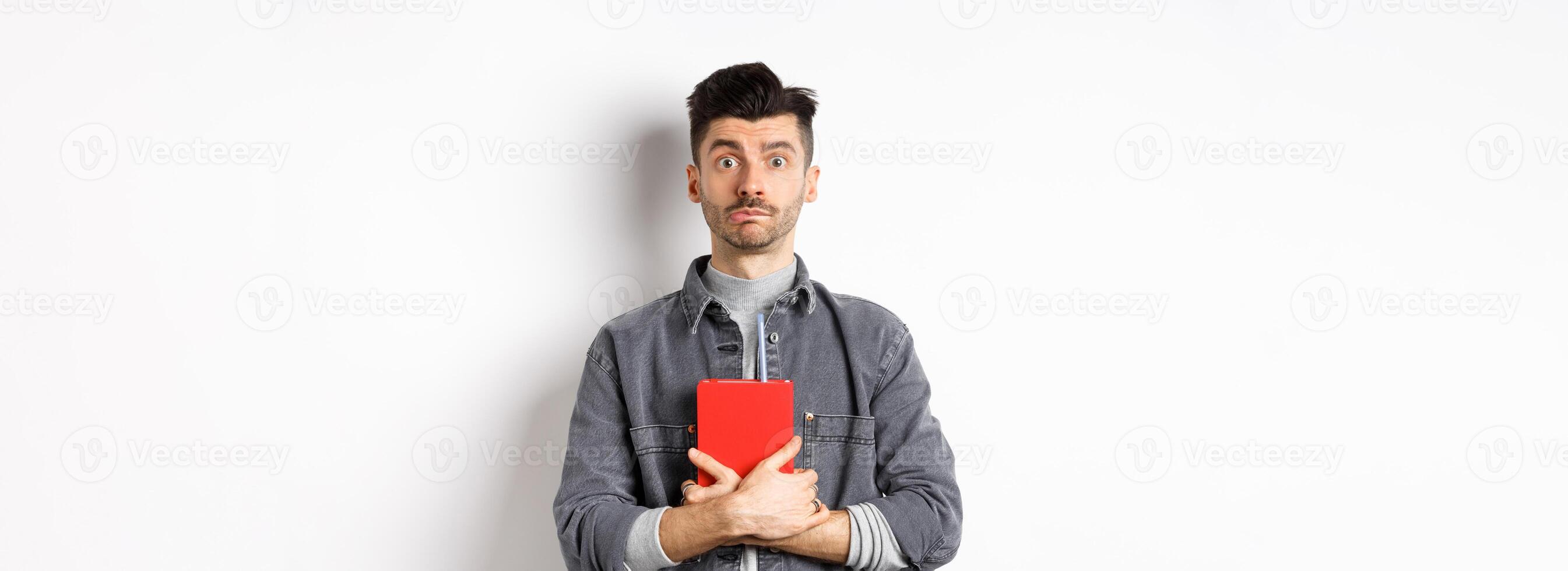 Cute and shy guy hugging his diary, holding red journal and look modest at camera, standing against white background photo