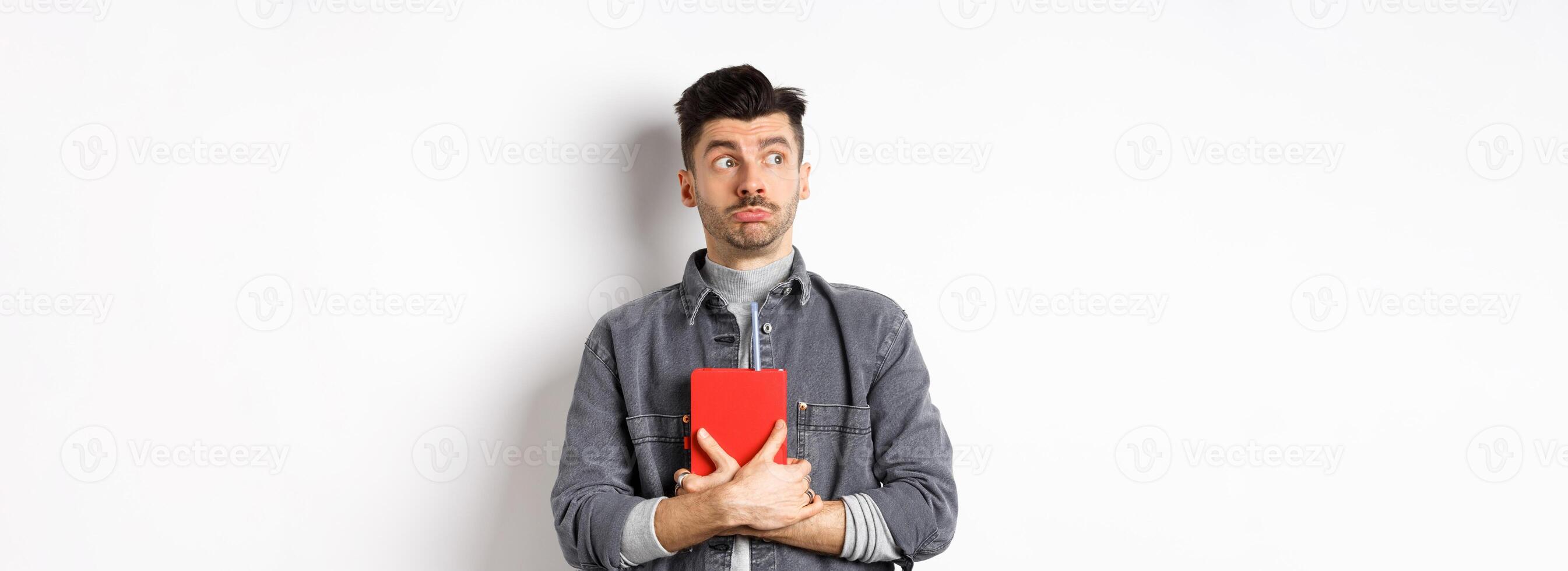Silly young man looking aside at logo with timid face, holding his diary pressed to chest, carry journal, standing on white background photo
