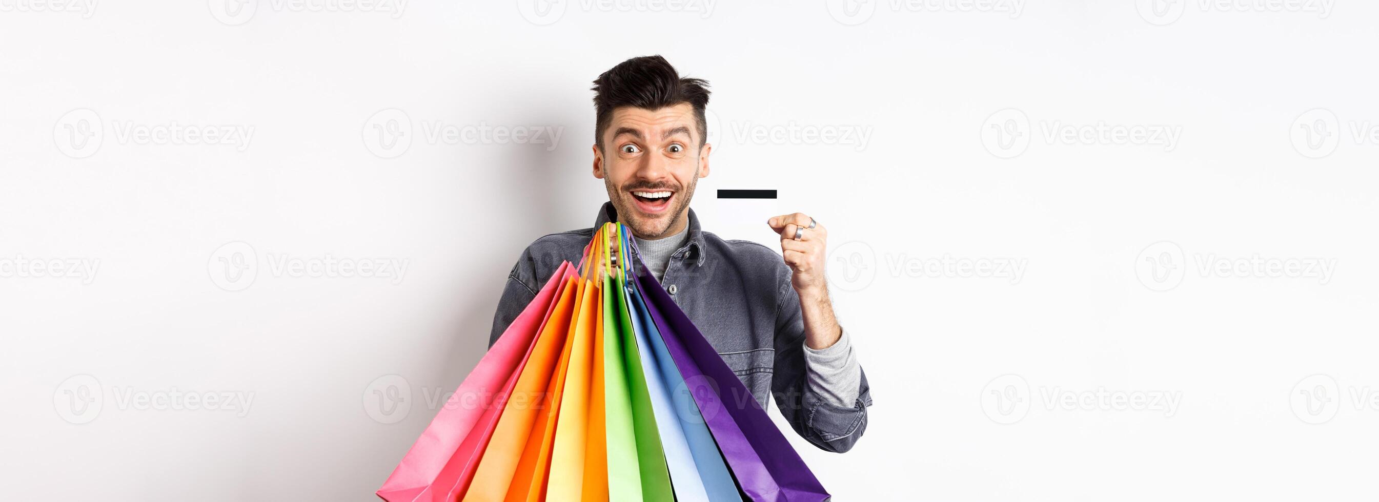 Happy guy shopaholic holding colorful shopping bags and showing plastic credit card, smiling excited, standing white background photo