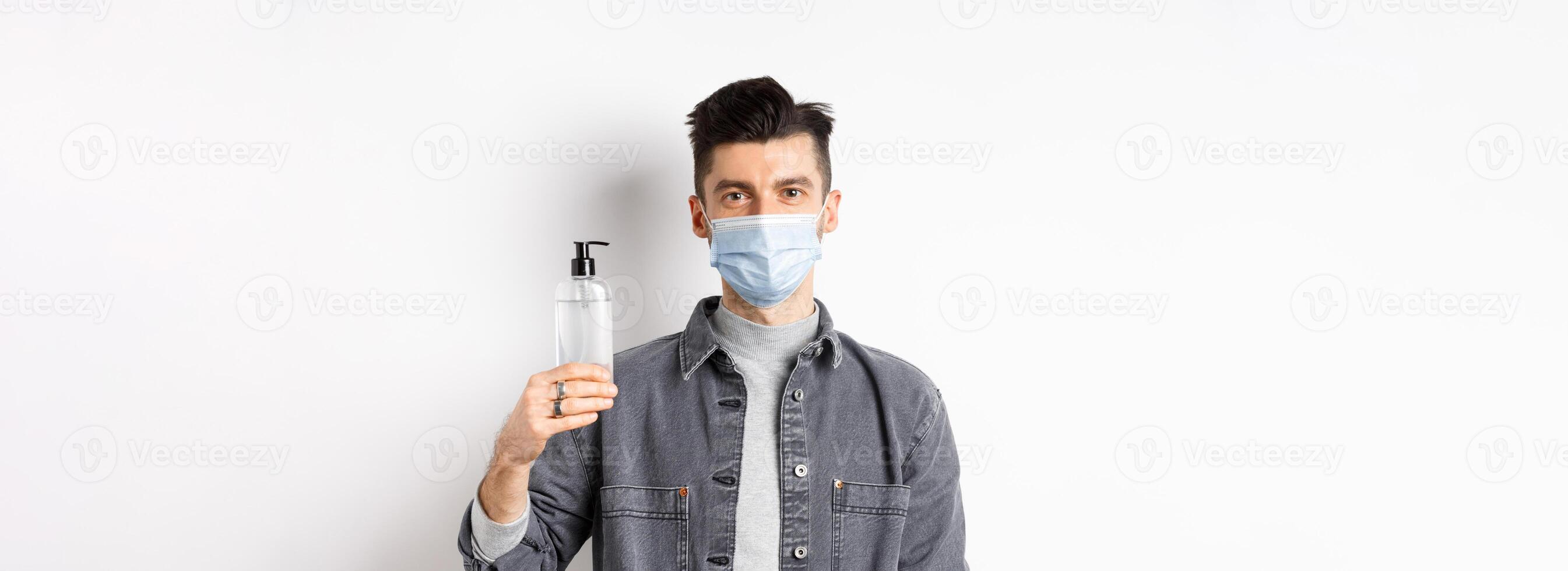 Healthy people and covid-19 concept. Young man in medical mask use preventive measures from coronavirus, showing bottle of hand sanitizer, standing against white background photo