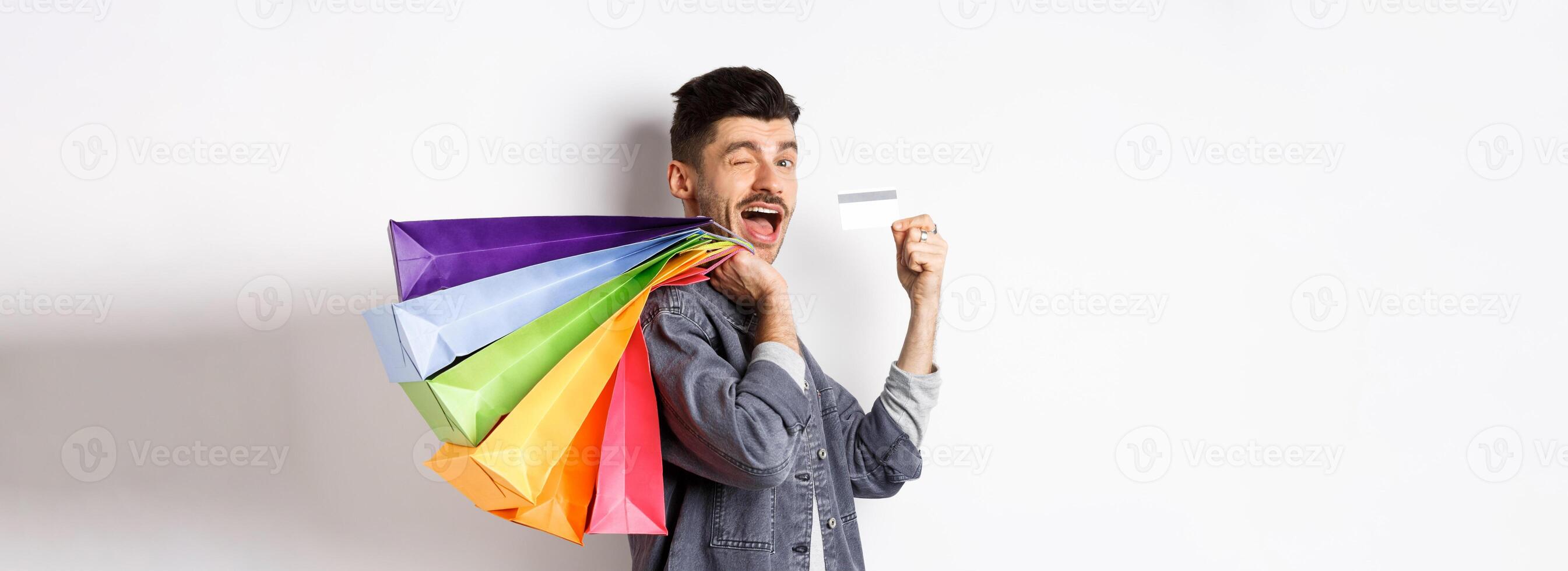 Happy shopper holding shopping bags over shoulder and showing plastic credit card, paying contactless, standing on white background photo