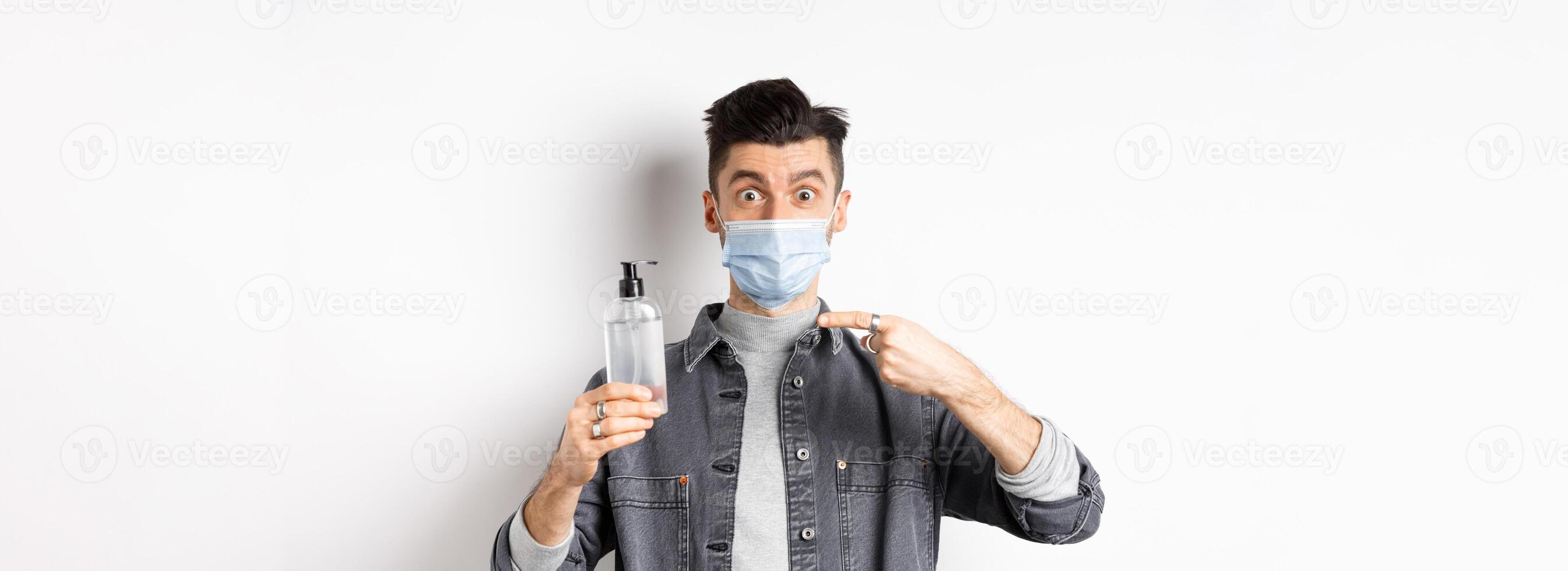 Health, covid and pandemic concept. Stylish guy in medical mask pointing at bottle with hand sanitizer, showing antiseptic, standing on white background photo