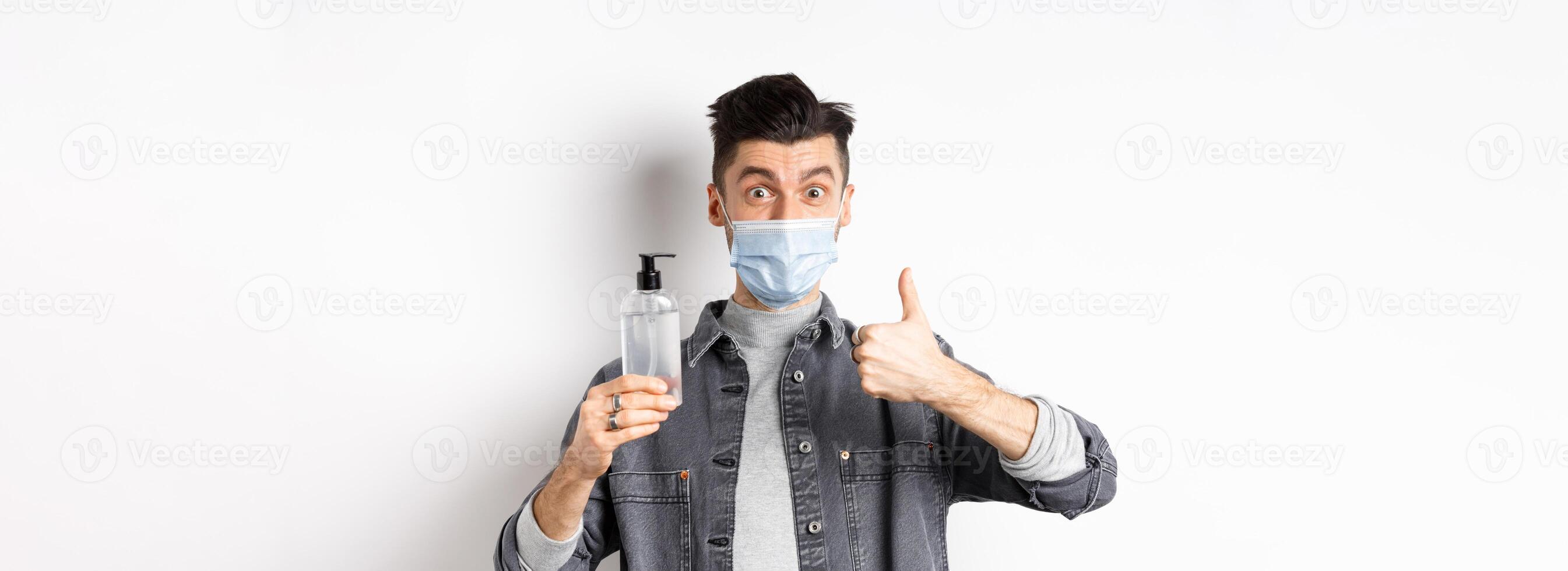 Healthy people and covid-19 concept. Excited man in sterile medical mask holding bottle of good hand sanitizer, show thumb up, recommend antiseptic, standing against white background photo