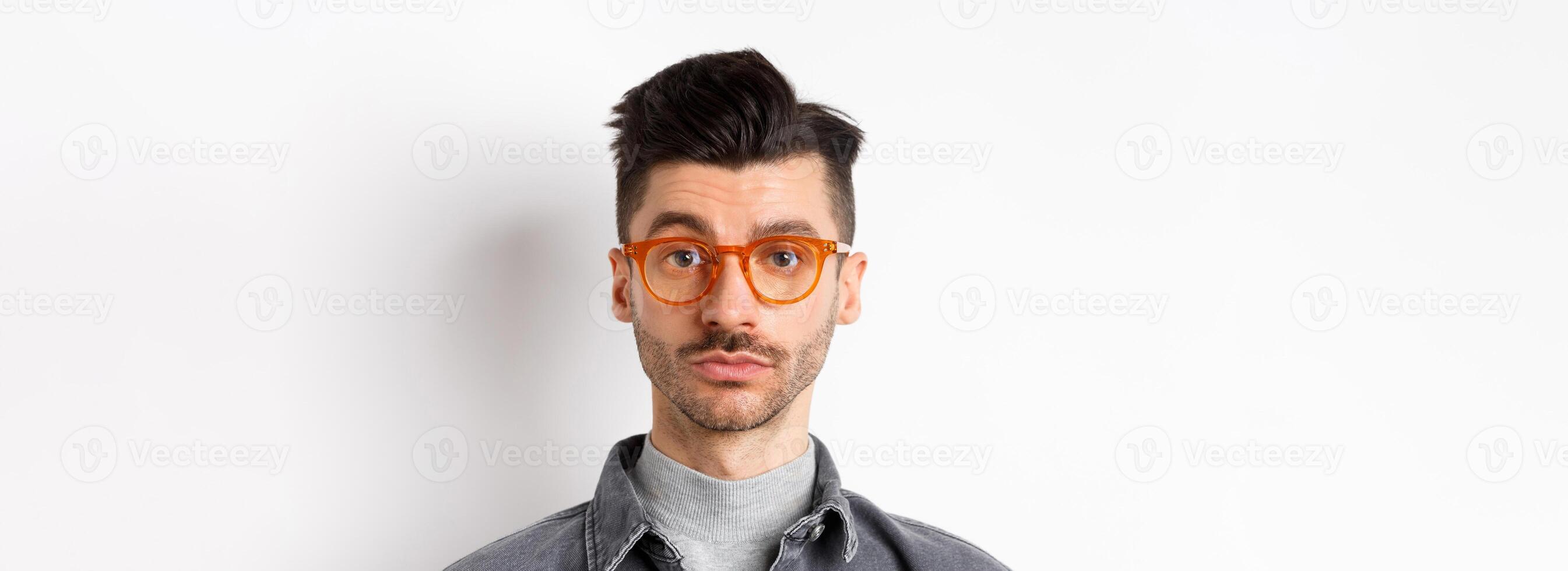 Attractive brunette man with moustache, wearing stylish glasses and look serious at camera, no emotion pokerface, standing against white background photo