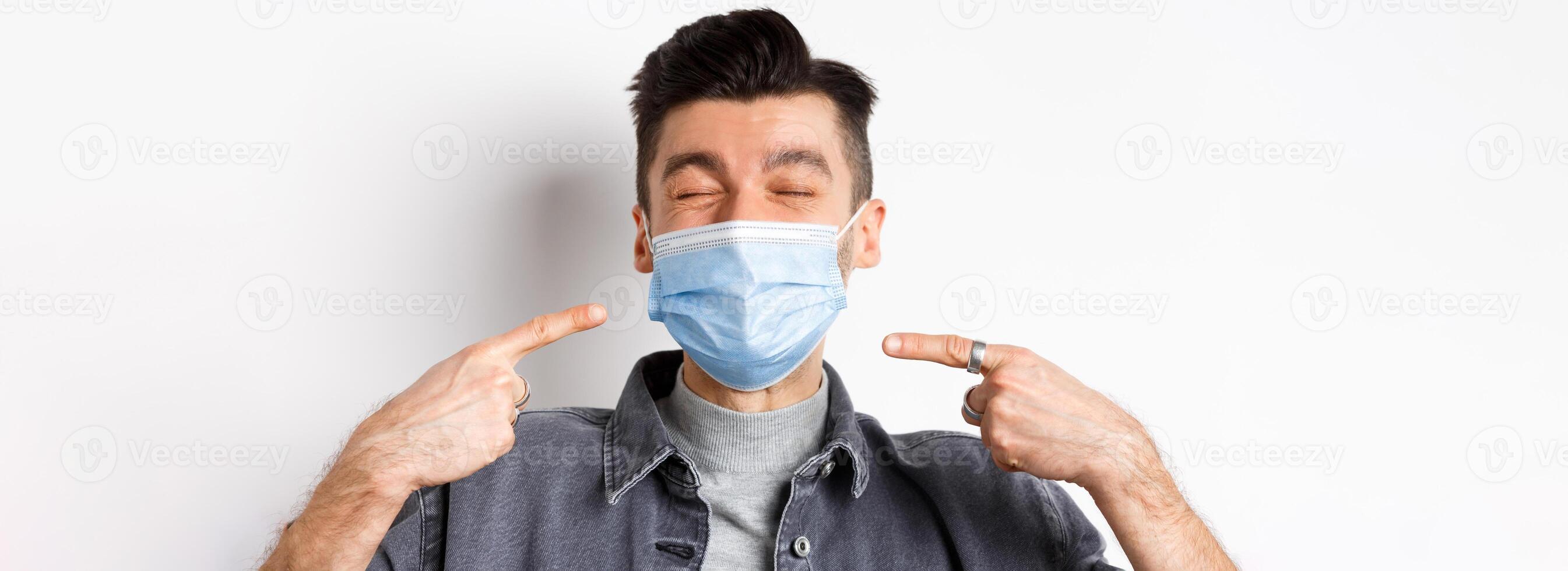 Covid-19, health and lifestyle concept. Cheerful smiling man pointing at sterile medical mask, protect himself from corona outbreak, standing on white background photo