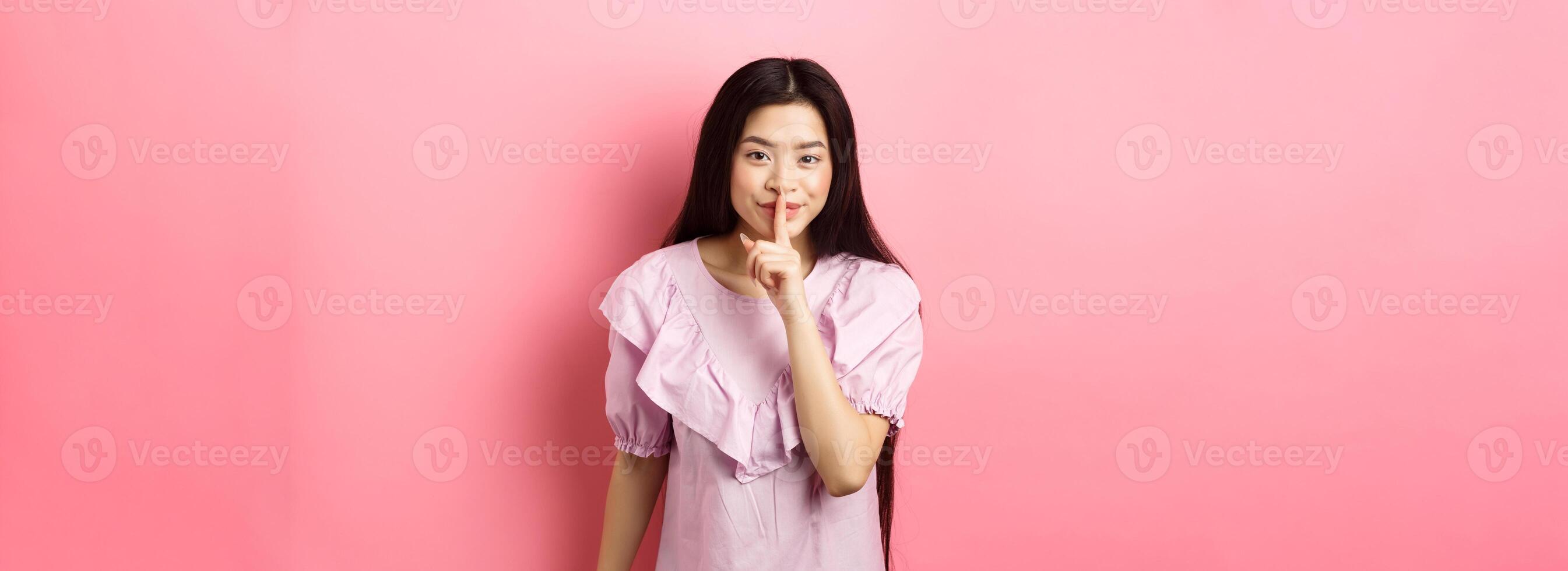 Cute asian girl hiding secret, hushing with finger pressed to lips and smiling, asking to keep quiet, standing in dress on pink background photo