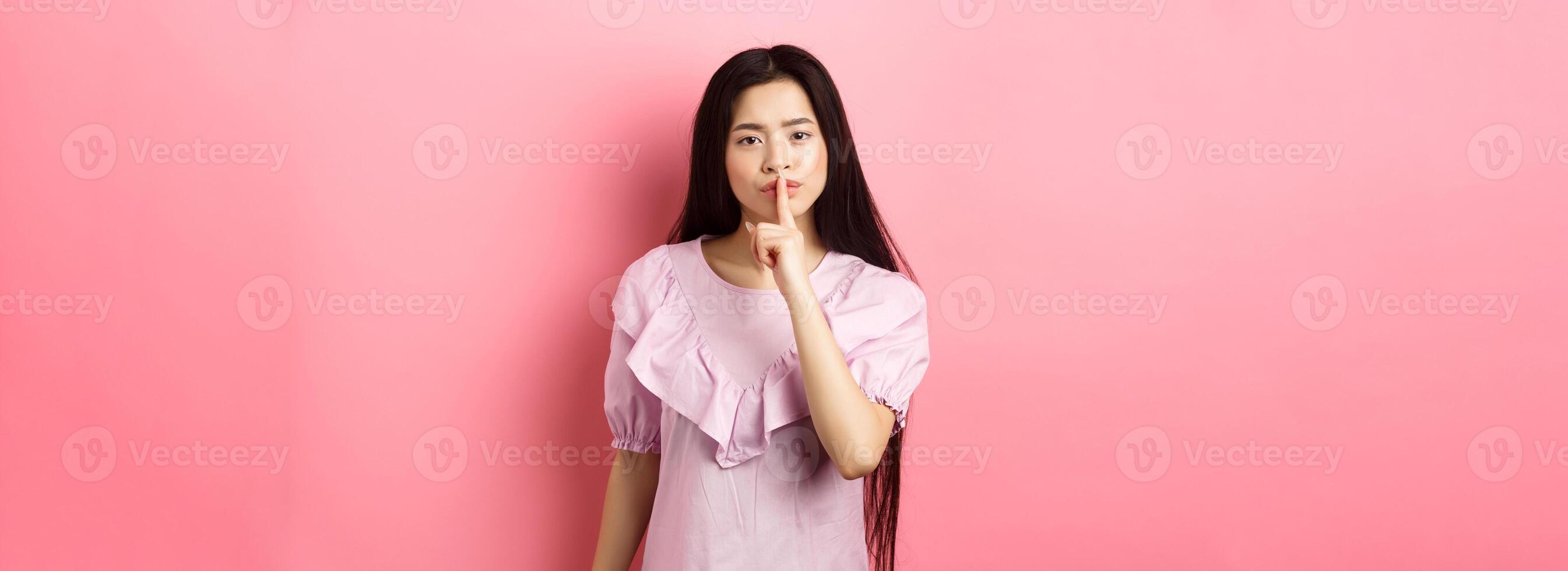 Cute asian girl tell to be quiet, scolding loud person, showing shush sign with finger pressed to lips, standing in dress on pink background photo