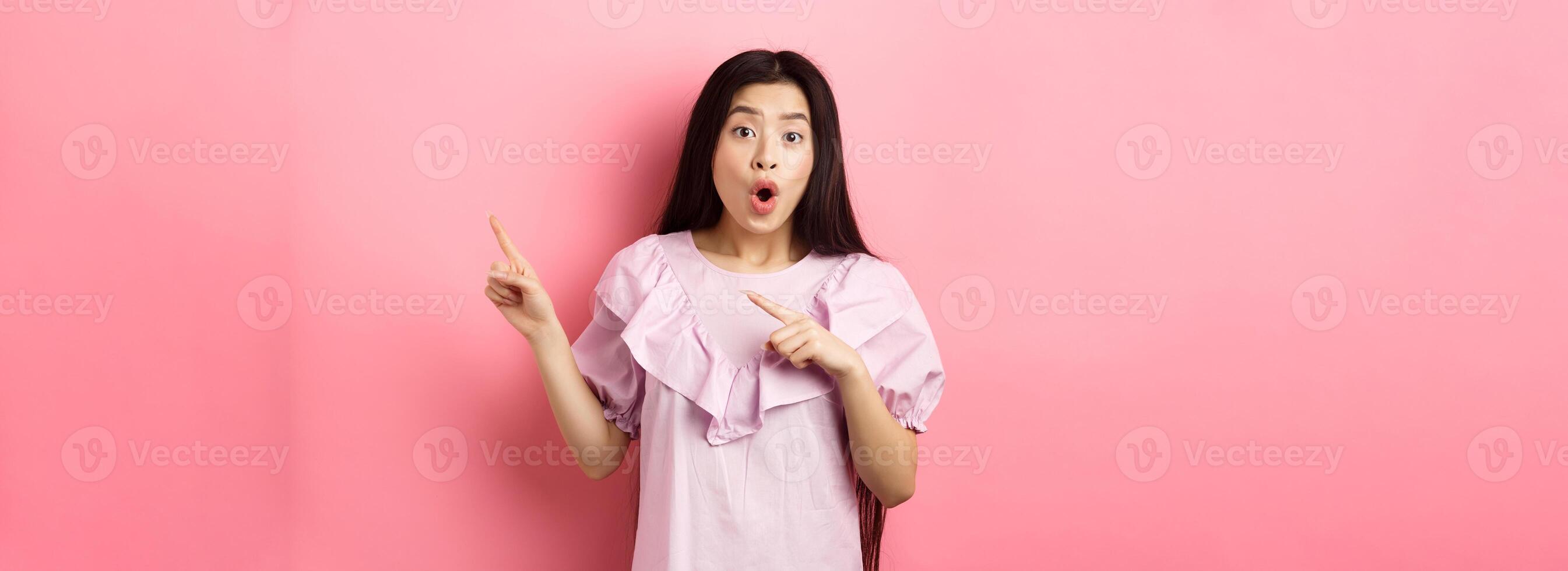 Excited asian woman look amazed, pointing right and gasping fascinated, checking out advertisement, standing on pink background photo