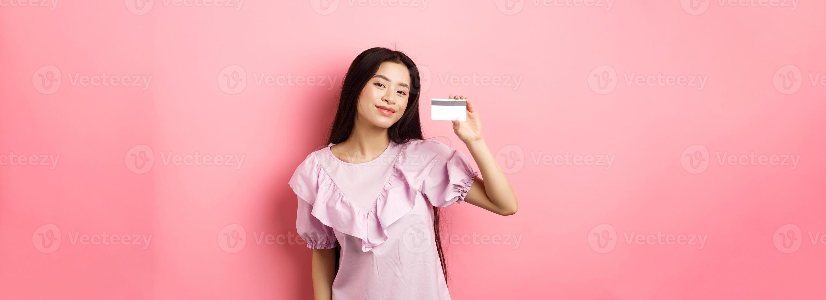 Shopping. Young korean girl in dress showing credit card, smiling satisfied at camera, standing on pink background photo
