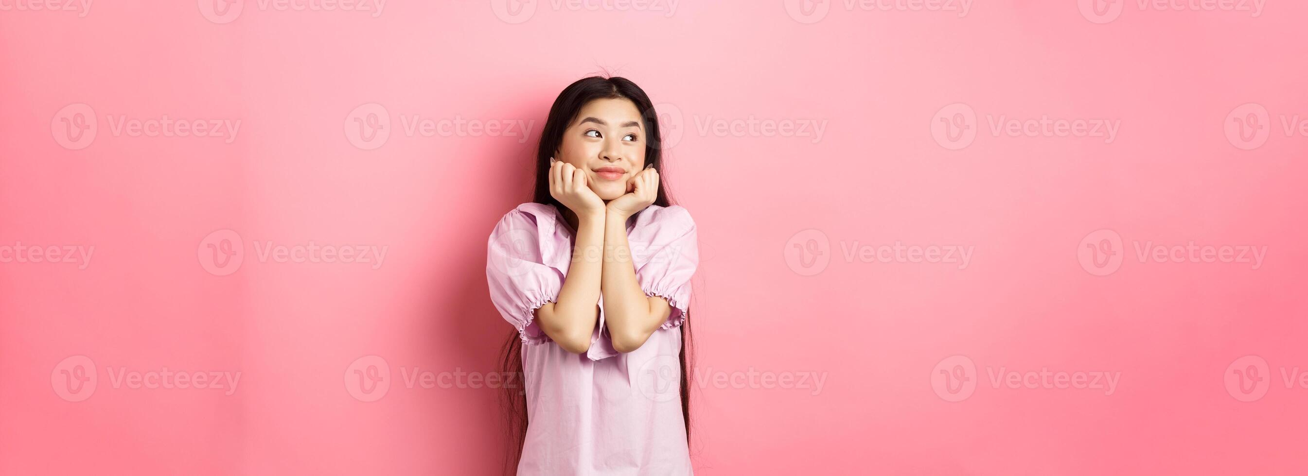 Dreamy romantic girl looking aside at logo and smiling, thinking of something beautiful, standing on pink background photo