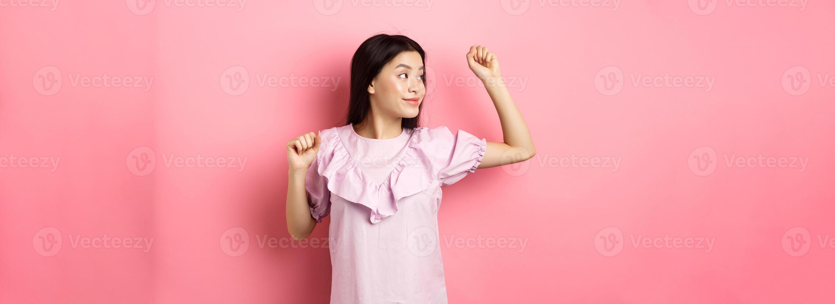 Carefree asian girl dancing, raising hand up and looking left at logo, standing in romantic dress against pink background photo