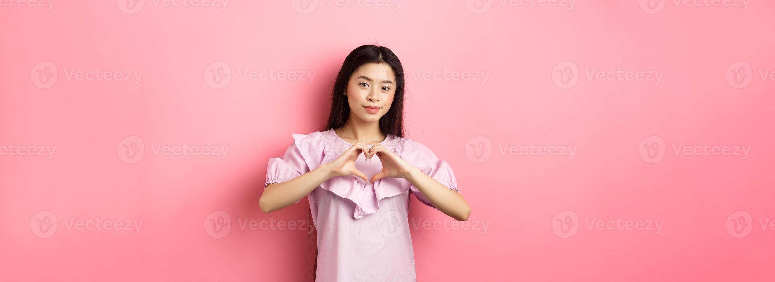 Valentines day concept. Beautiul japanese teen girl show heart gesture and smiling cute, express love and sympathy to lover, standing in romantic dress on pink background photo