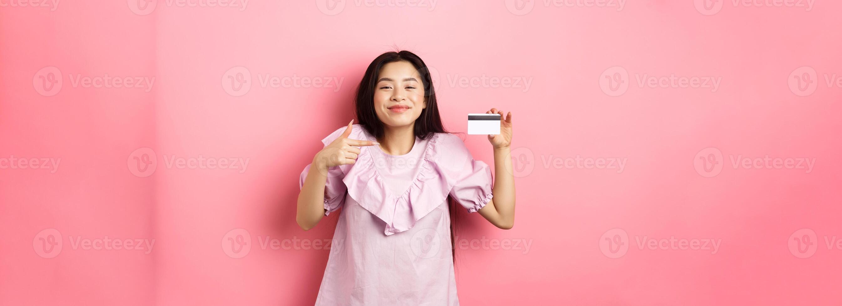 Cheerful and cute asian woman smiling satisfied, showing plastic credit card, standing in dress against pink background photo