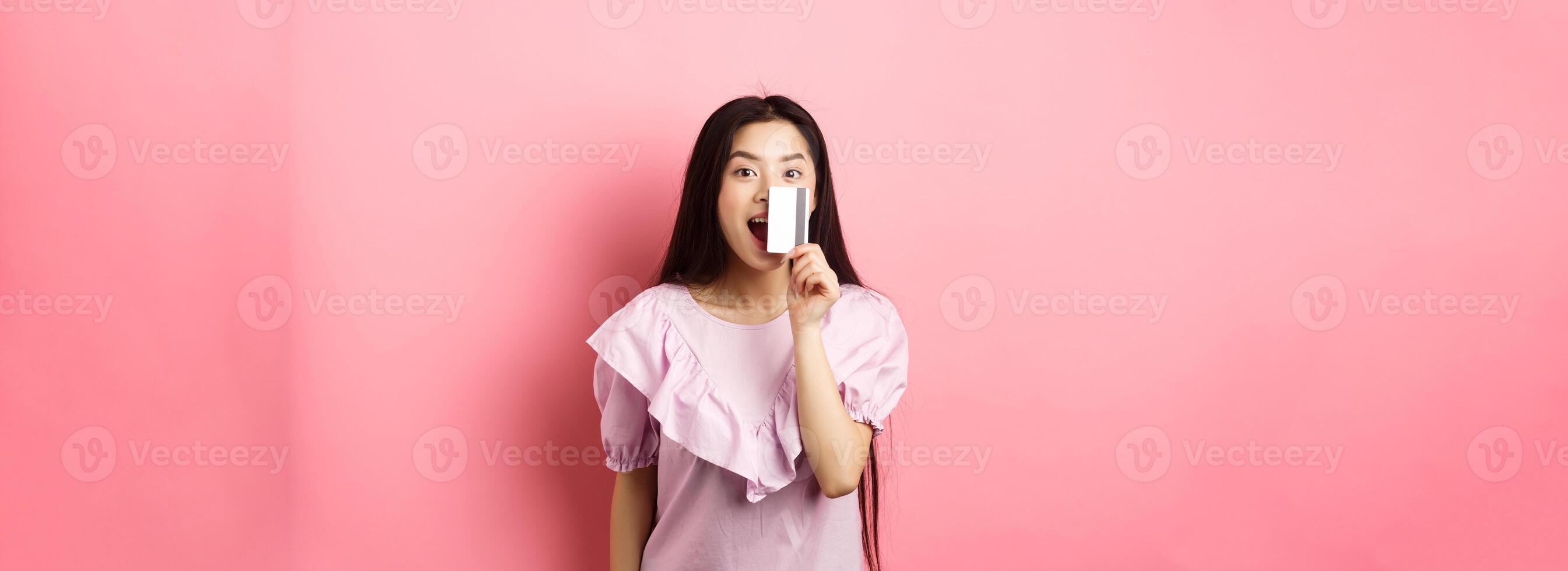 Excited asian girl showing plastic credit card and smiling, going on shopping, standing against pink background photo