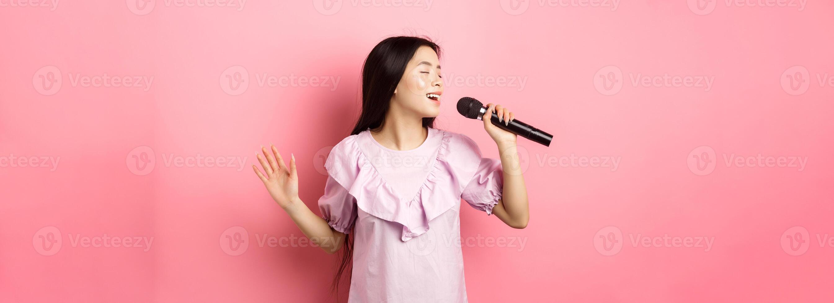 Beautiful asian girl perform song, singing in microphone and smiling romantic, standing in dress against pink background photo