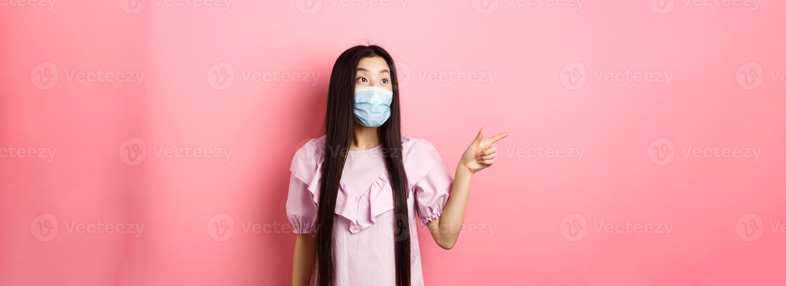 Coronavirus, quarantine and lifestyle concept. Excited asian teen girl in face mask pointing, looking left at logo with amazed face, checking out promotion, white background photo