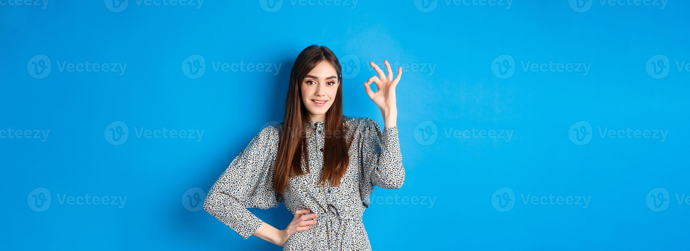 Confident young woman in dress showing okay sign and smiling, no problem gesture, assure everything good, standing on blue background photo