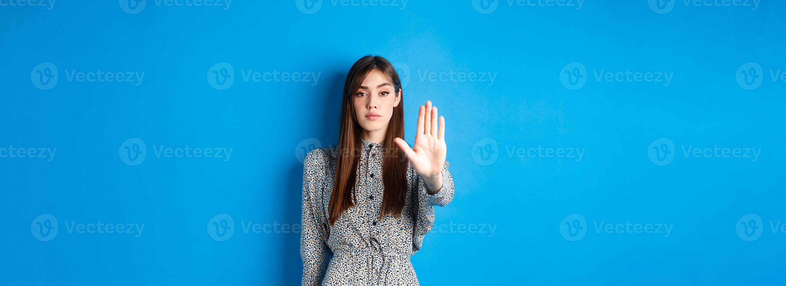 Serious and confident woman with long hair and dress, stretch out hand and say no, make stop gesture, prohibit bad action, standing on blue background photo
