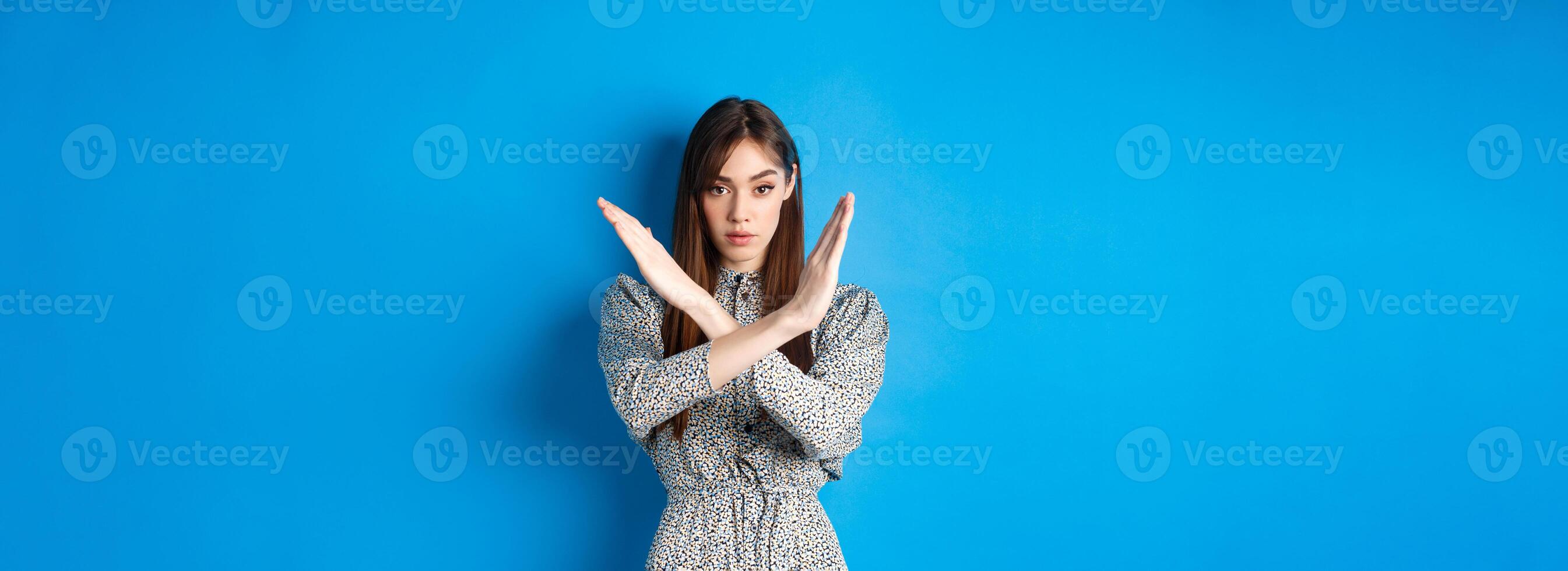 Serious caucasian woman in dress tell no, show cross stop gesture and looking confident, making warning sign, prohibit something bad, standing on blue background photo