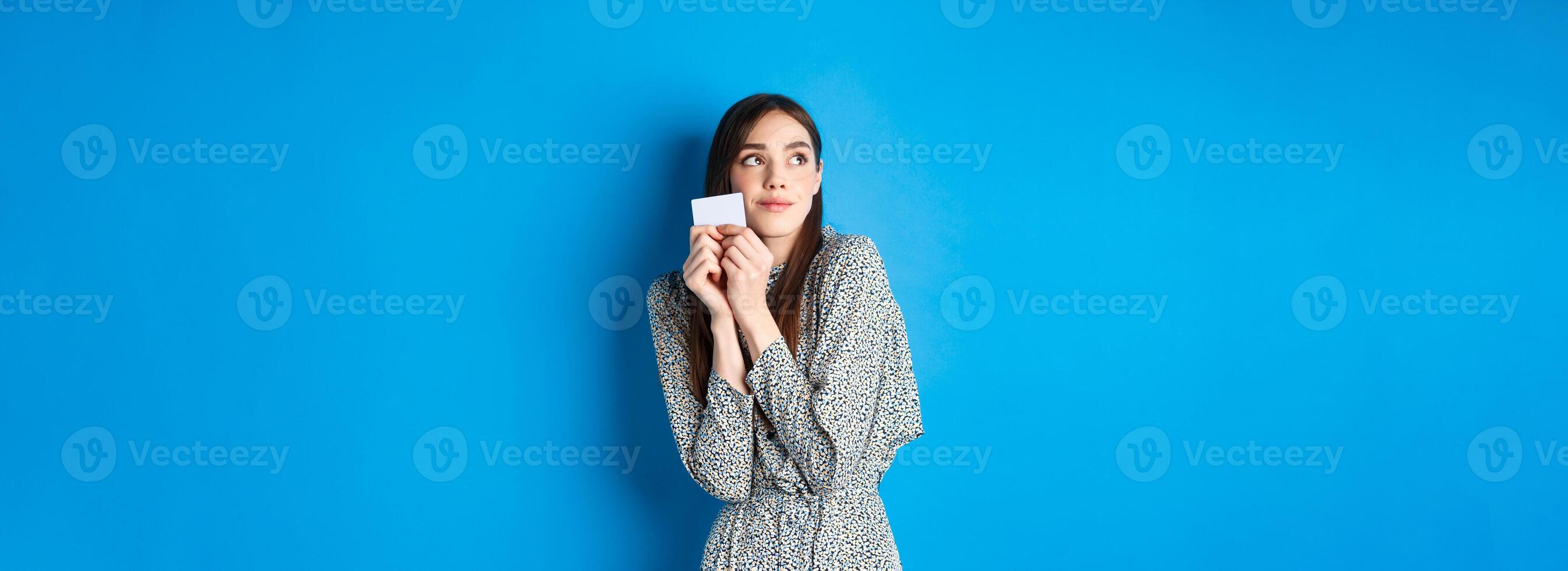 Romantic cute girl in dress dreaming of shopping, holding credit card and looking at logo with tender smile, standing on blue background photo