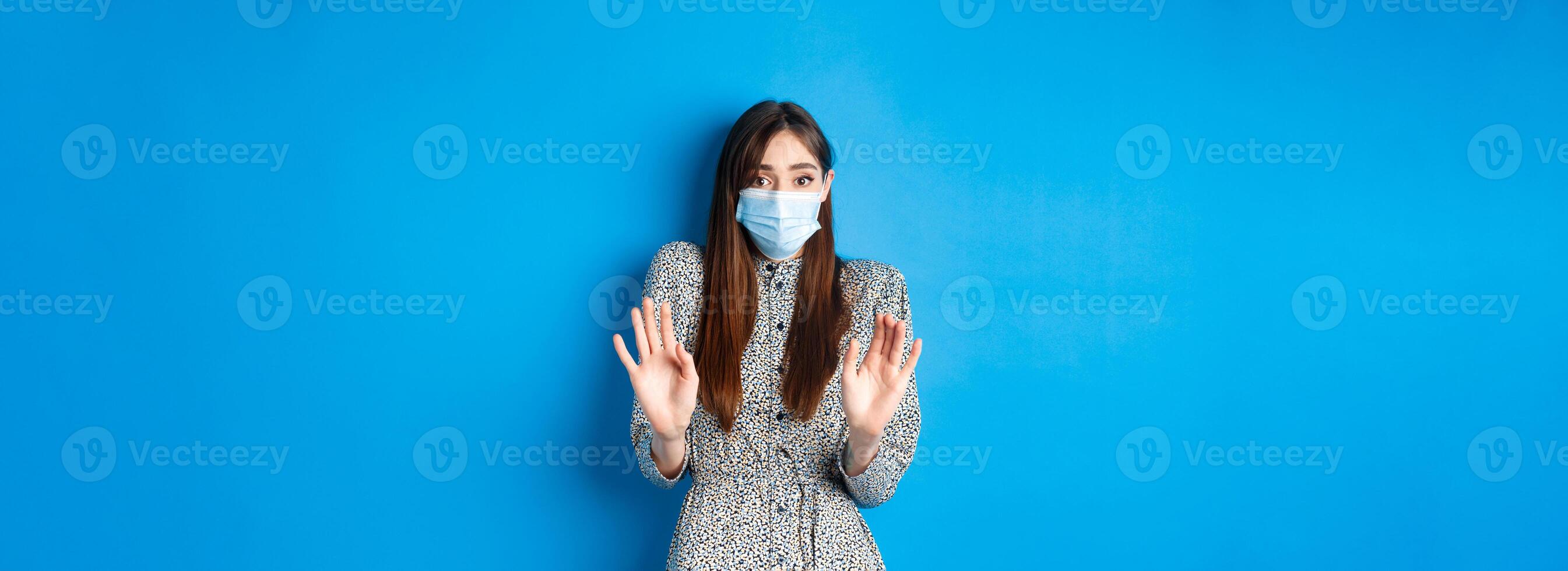 Covid-19, pandemic lifestyle concept. No thank you. Reluctant and worried girl in medical mask asking non-masker to stay away, showing block gesture, tell stop, blue background photo