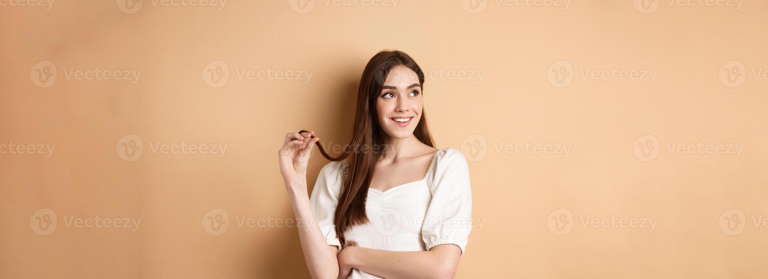 Romantic pensive girl thinking, looking left at empty space and playing with hair strand, smiling dreamy, standing in white dress on beige background photo