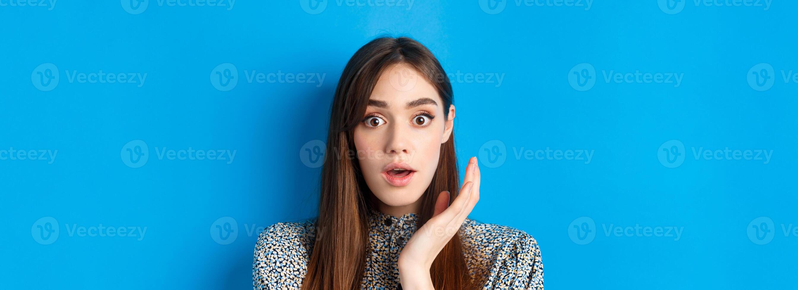 Candid girl with long hair and makeup, looking shocked and surprised, open mouth and stare speechless at camera, standing on blue background photo