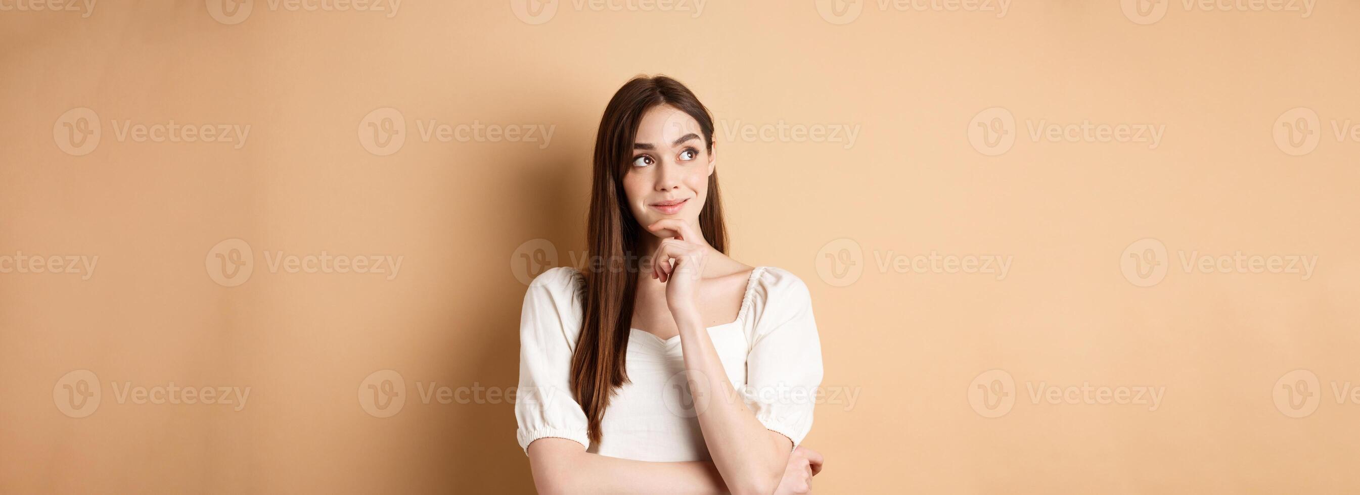 Very interesting. Pensive cute girl thinking and looking at upper left corner logo, smiling pleased, having good idea, standing on beige background photo
