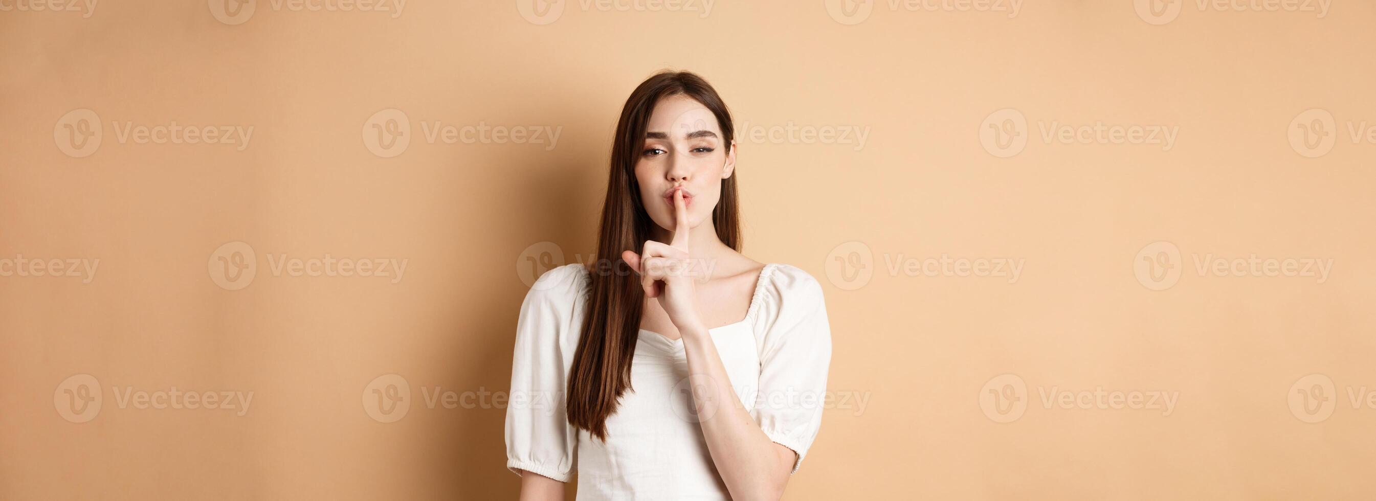 Sensual cute girl hiding secret, showing hush gesture, make shhh sound with finger on lips, stay quiet sign, standing on beige background photo