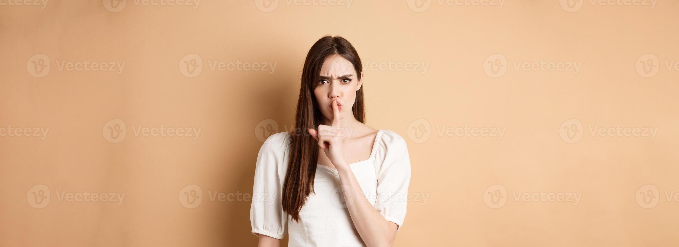 Angry young girl scolding bad behavious, shushing and frowning mad, tell to be quiet, looking displeased, standing on beige background photo