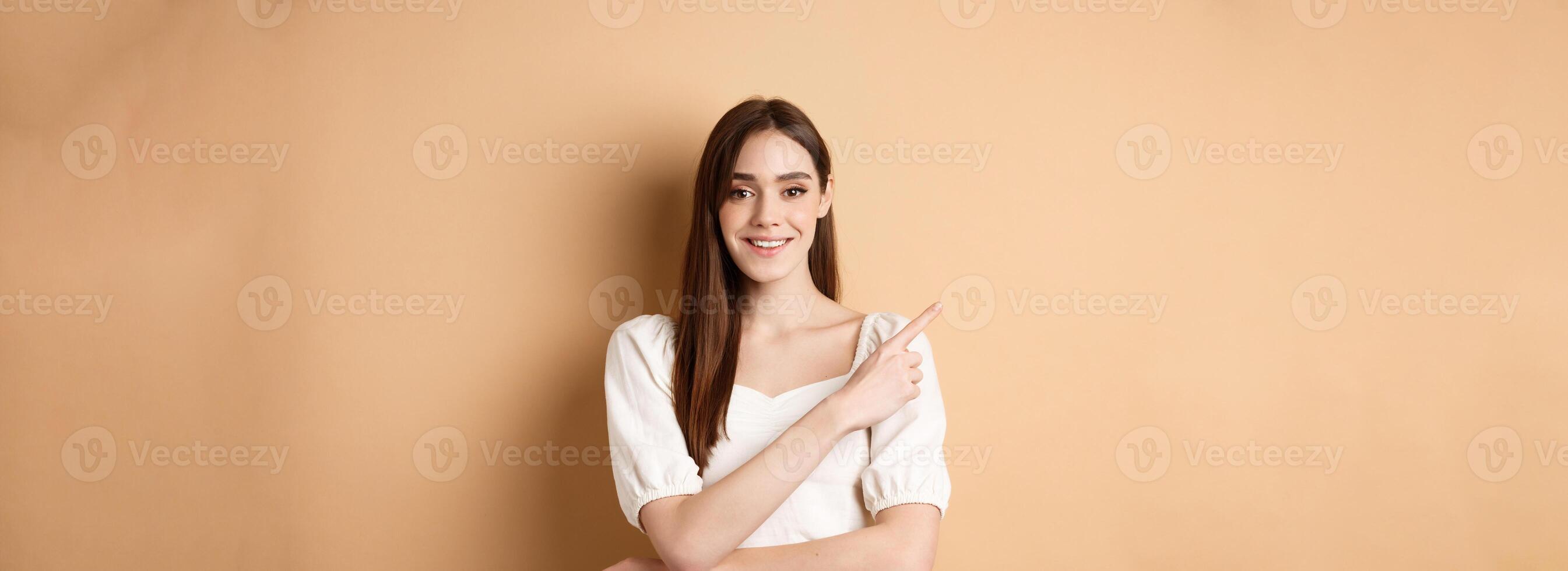 Young cheerful female model in romantic dress pointing finger at upper left corner, smiling pleased at camera, showing her choice, recommending product, beige background photo