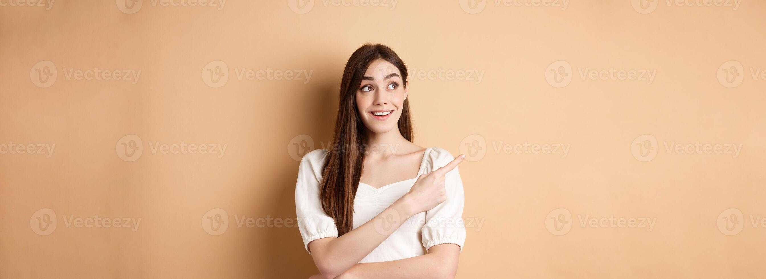 Cheerful tender girl in white dress checking out promo offer, looking and pointing at upper left corner with dreamy smile, standing on beige background photo