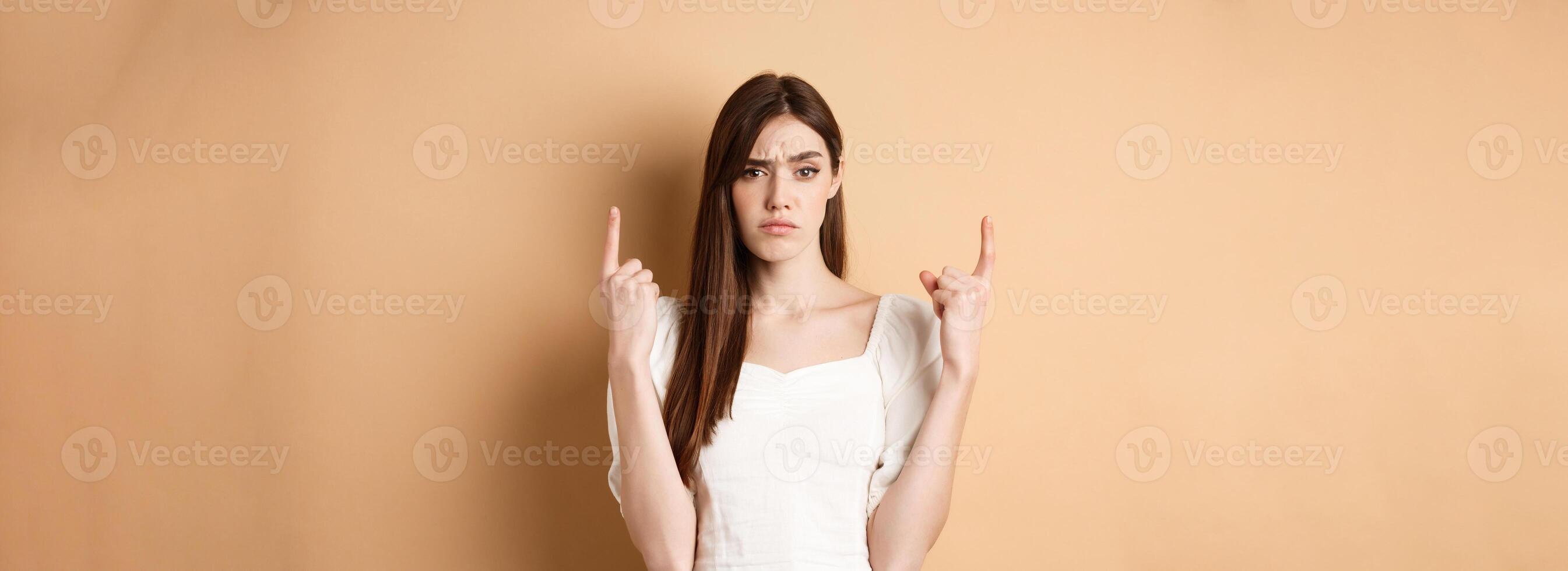 Skeptical woman frowning and pointing fingers up, having doubts about product, standing on beige background photo