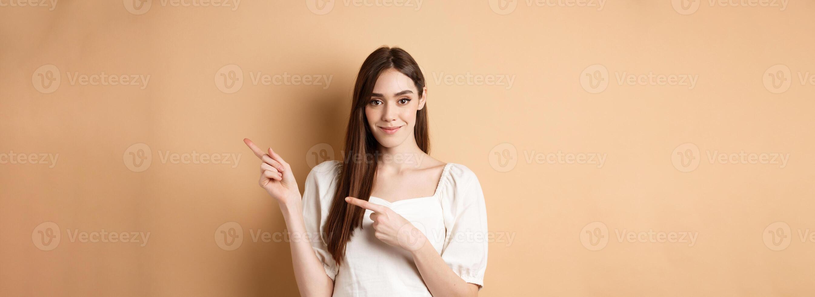 Beautiful young woman in dress pointing fingers right, showing logo and looking tender at camera, beige background photo