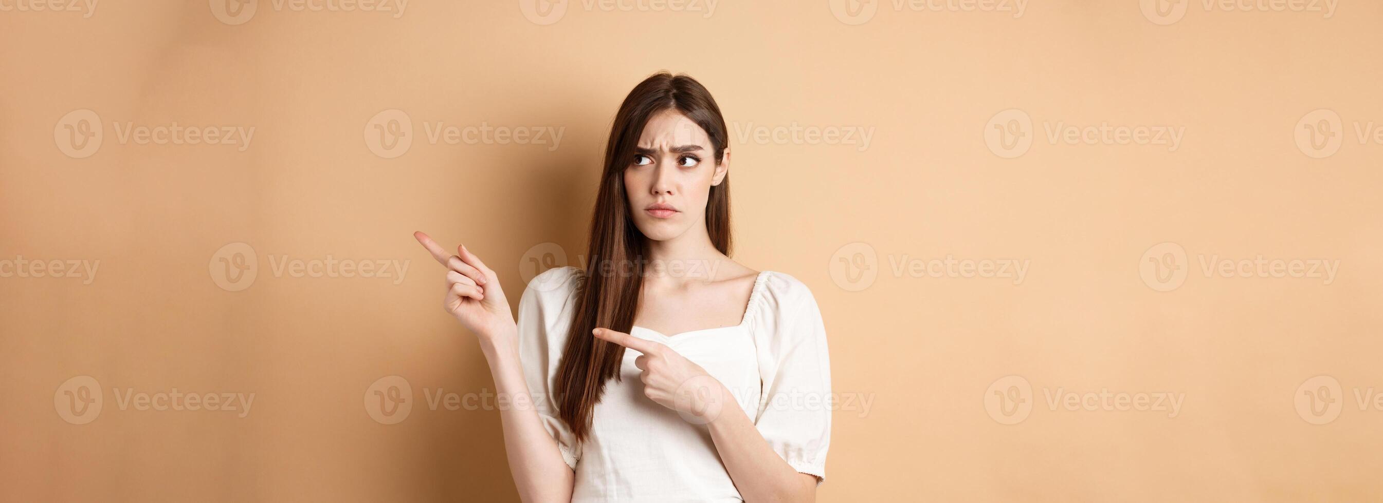 Hesitant young woman look unsure, pointing fingers right while making decision, frowning with puzzled face, standing on beige background photo