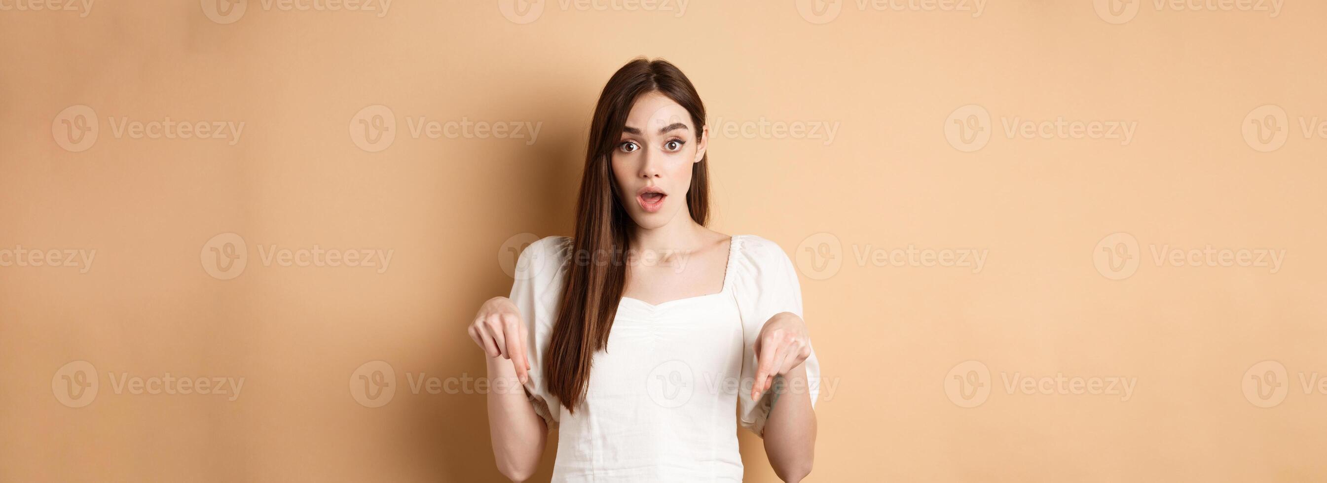 Wow look there. Amazed young woman in white dress pointing fingers down, standing in awe with dropped jaw and popped eyes against beige background photo