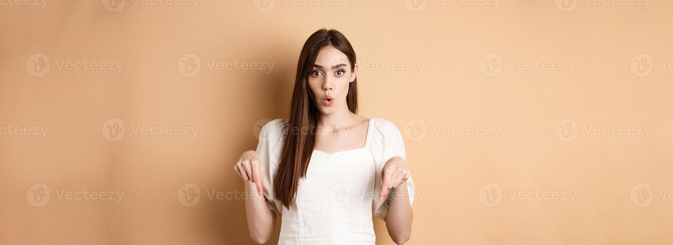 Wow amazing. Impressed young woman look astonished by product, pointing fingers down at logo, standing on beige background photo