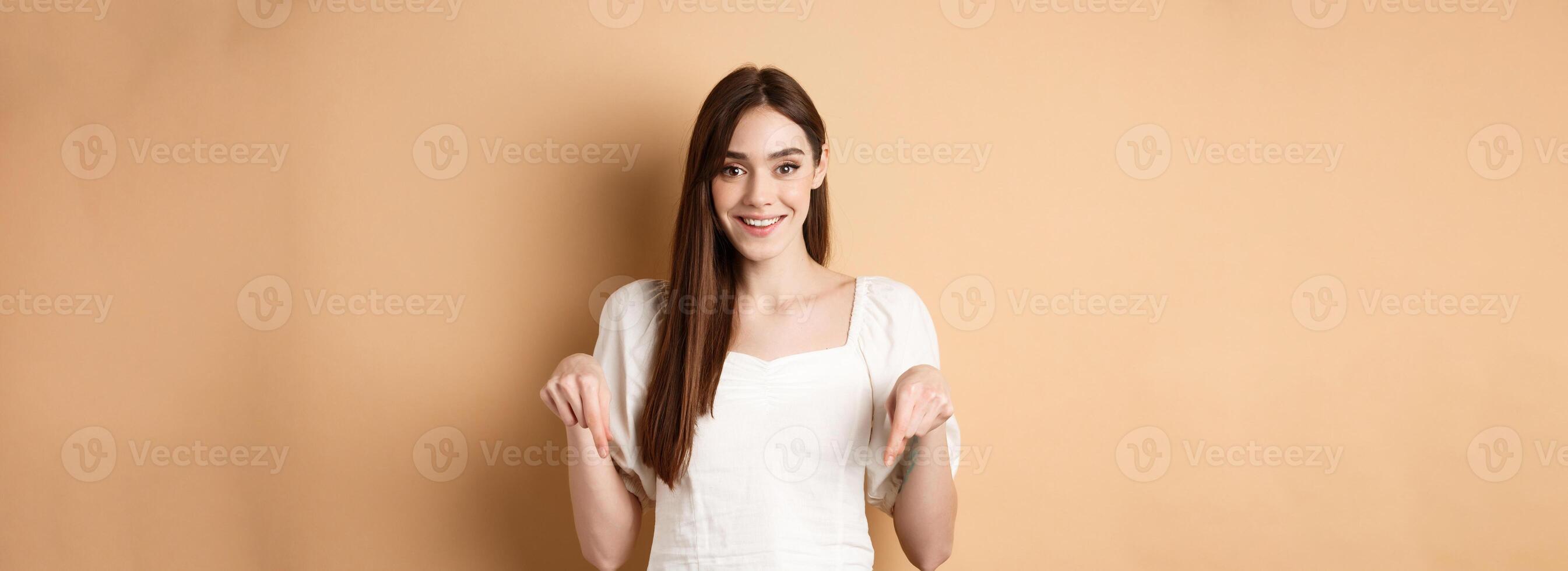 Cheerful young girl with lovely smile, pointing fingers down, making announcement or inviting click link, showing logo, beige background photo